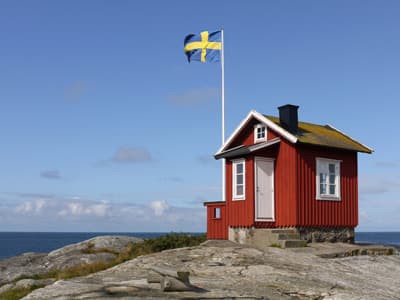 Ein einsames Schwedenhaus steht mit gehisster Flagge auf einem Felsen am Meer.