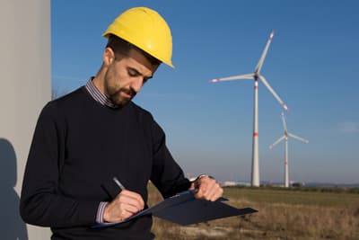 Ein Ingenieur mit gelbem Schutzhelm notiert sich etwas vor einer Windkraftanlage.