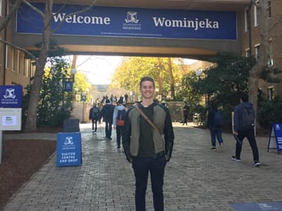 Ein junger Mann auf einem Campus vor einem Welcome-Schild