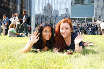 Zwei Studentinnen liegen im Gras vor der Hochschule und winken in die Kamera.