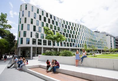 Studierende sitzen vor einem modernen länglichen Gebäude mit grüner Wiese