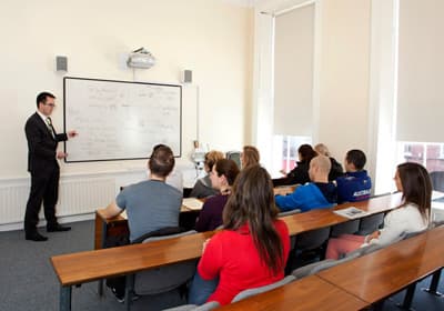 Kleine Gruppe von Studenten in einem Seminar des Dorset Colleges, der Dozent trägt einen Anzug und präsentiert etwas auf einem Whiteboard