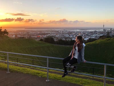 Eine junge Frau sitzt auf einem Geländer auf einem Berg und schaut in den Sonnenuntergang. Im Tal im Hintergrund sieht man die Stadt Auckland.