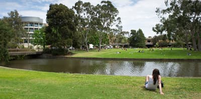 Ein Campus mit großen Grünflächen auf denen Studierende sitzen werden von einem Fluss geteilt