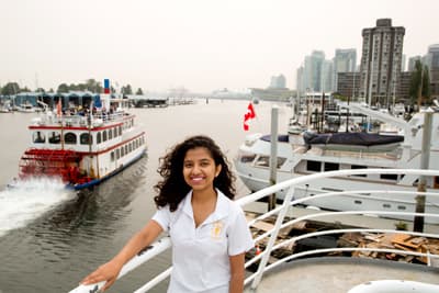 Tourism Management Studentin an Deck eines Schiffes. Im Hintergrund fährt ein Raddampfer vorbei und man sieht die Hochhäuser der Innenstadt Vancouvers.