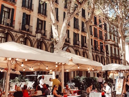 Sommerliche Caféterasse mit Lichterkette im Sonnenschirm vor einer schönen Altbaufassade und unter Platanen in Barcelona.