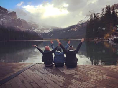 Drei Freundinnen mit Mützen sitzen an einem See mit Gebirgspanorama und jubeln