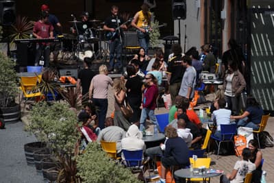 Gut besuchte Caféterasse, auf der Menschen in der Sonne sitzen, im Hintergrund spielt eine Band Livemusik