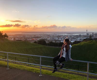 Studentin posiert auf einem Geländer am grünen Mount Eden. Im Hintergrund sieht man die Stadt Auckland aus der Ferne mit ihrem charakteristischen Sky Tower.