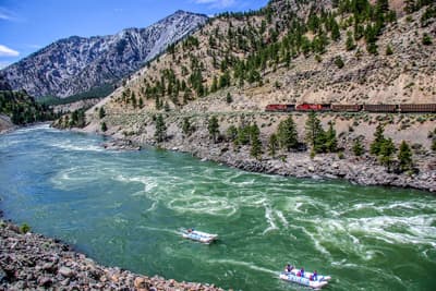 Reißender türkisfarbener Fluss, auf dem zwei Rafting-Boote fahren vor einer Berglandschaft mit Zug