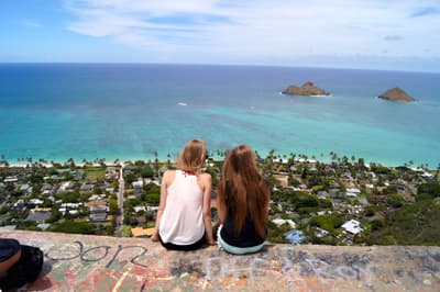 Zwei Studentinnen sitzen auf einem Aussichtspunkt mit Blick über die traumhafte Küste Honolulus
