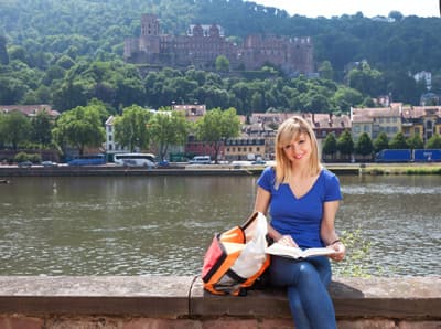 Eine Studierende sitzt in Heidelberg auf einer Mauer am Fluss.