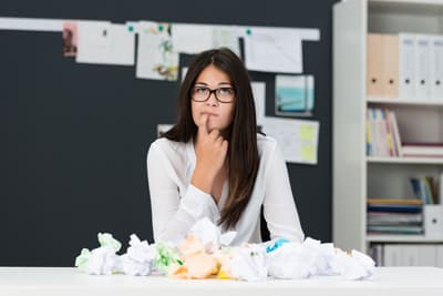 Eine junge Frau sitzt grübelnd vor einem Haufen zerknülltem Papier.