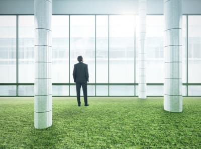 Ein Manager im Anzug steht in einem lichtdurchfluteten Gebäude auf einer grünen Rasenfläche und schaut nach draußen.