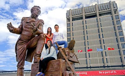 Drei Studenten auf einer Bronze-Statue von einem Mann vor einem Hochhaus