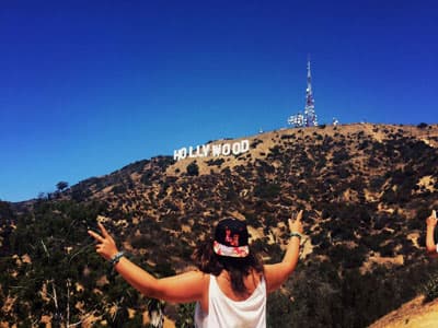 Eine Frau mit dem Rücken zu Kamera zeigt vor dem Hollywood Sign das Peacezeichen