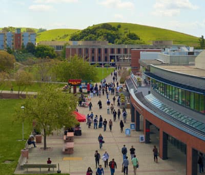 Eine Straße auf einem Universitätscampus mit Studierenden und Hügeln im Hintergrund