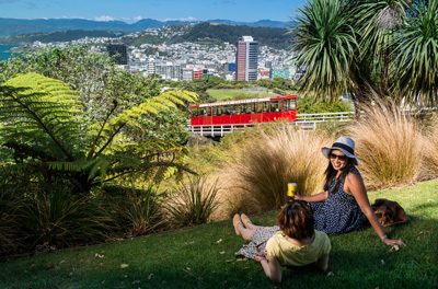 Zwei Freunde sitzen auf dem Rasen, während hinter ihnen die Seilbahn in Wellington den Berg hochfährt